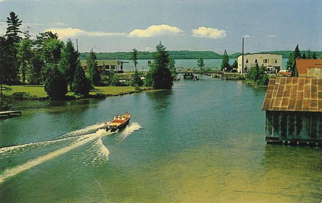 Early-1960s color postcard: a runabout heading down the Clam River toward the mouth, boathouse at right, the white store and docks ahead, Torch Lake beyond.