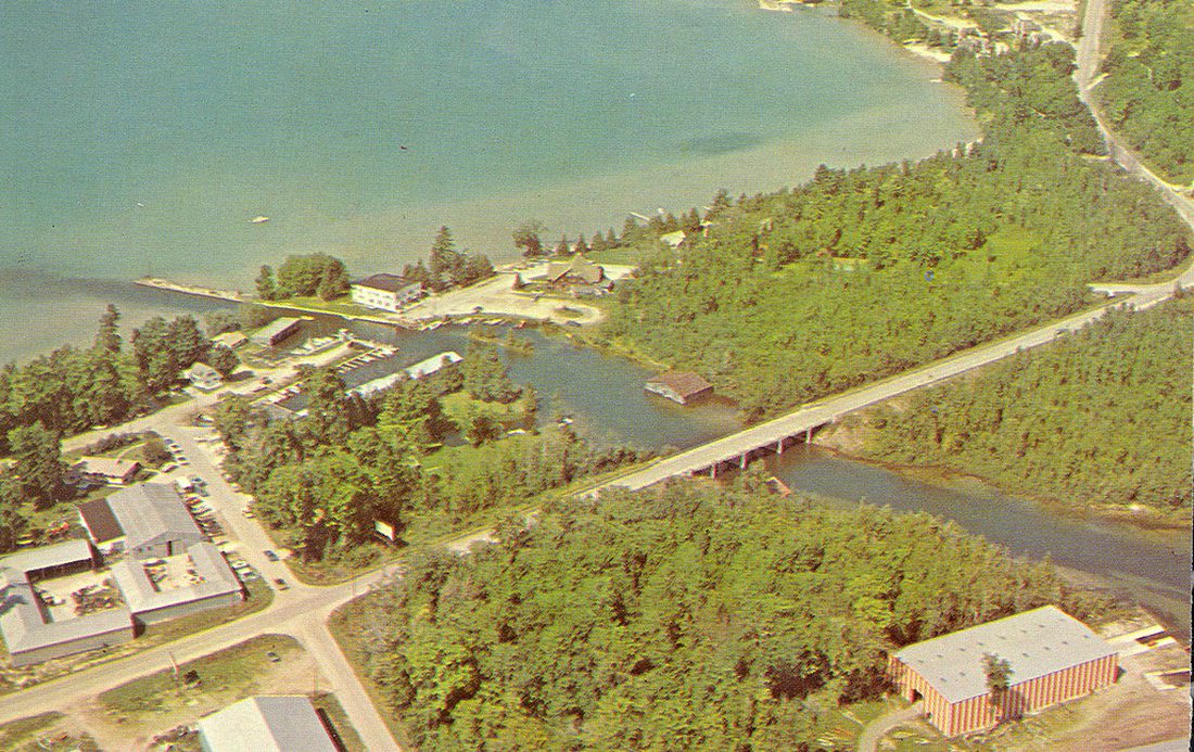 1960s-70s aerial photograph: the river mouth with bridge, a full small-craft marina, and along the east shore a sandy swimming beach with a low pavilion, picnic tables and parking.